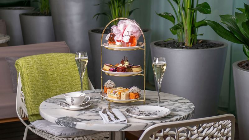 Discover the Mad Hatter's Afternoon Tea at Sanderson. Credit: Sanderson. Image courtesy of Sanderson. A photo of two chairs at a round table with plates and a tiered afternoon tea display featuring Alice in Wonderland-themed cakes and two glasses of bubbles