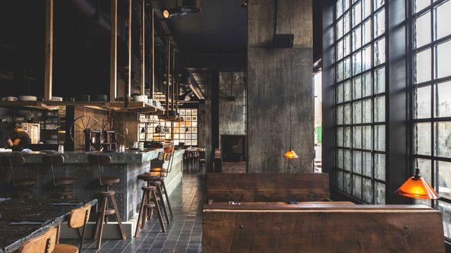 A moody restaurant with high windows brown tables and a long grey bar.