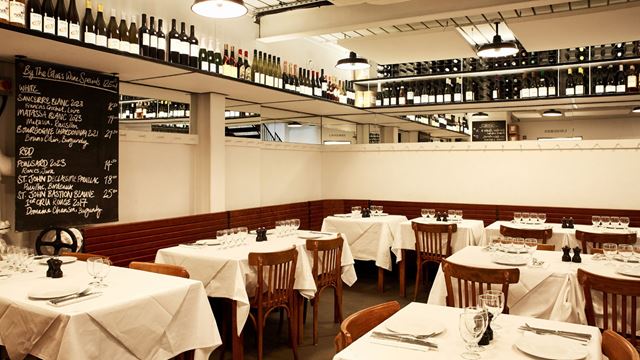 A restaurant with a chalkboard menu, white walls, a high shelf lined with wine bottles, and square tables covered with white tablecloths.