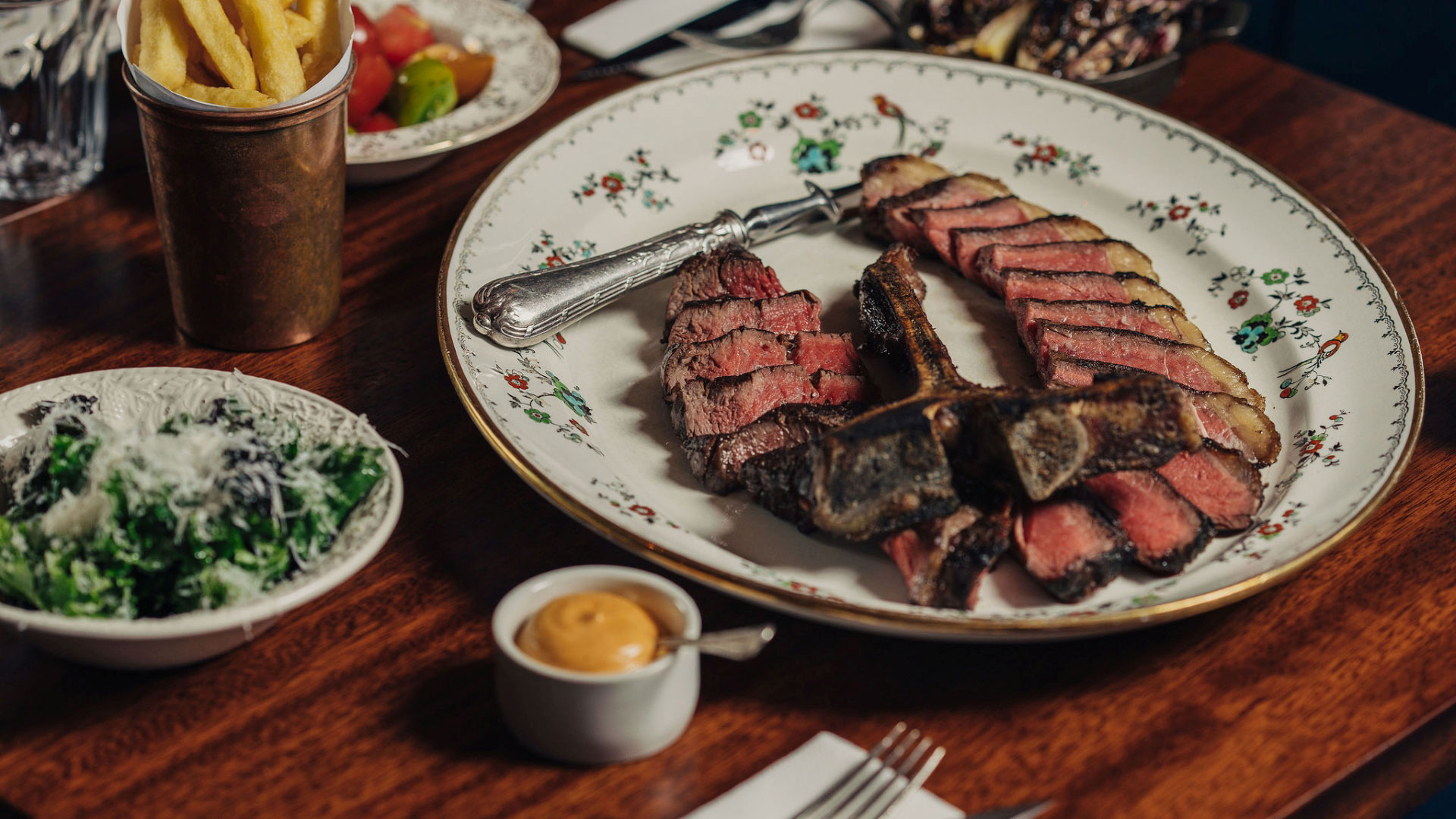 A photo of a vintage dish with a perfectly cooked T bone steak thinly sliced on top, with sides of fries and salad on a brown wood table at Blacklock