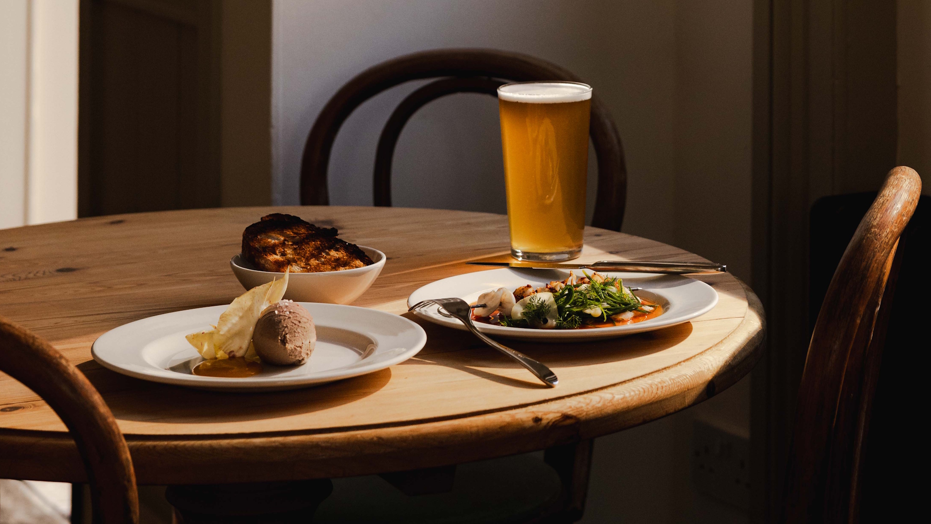 Wooden table and chairs with a pint of beer, an elegant seafood plate, some bread in a basket and a chocolate mousse dessert.