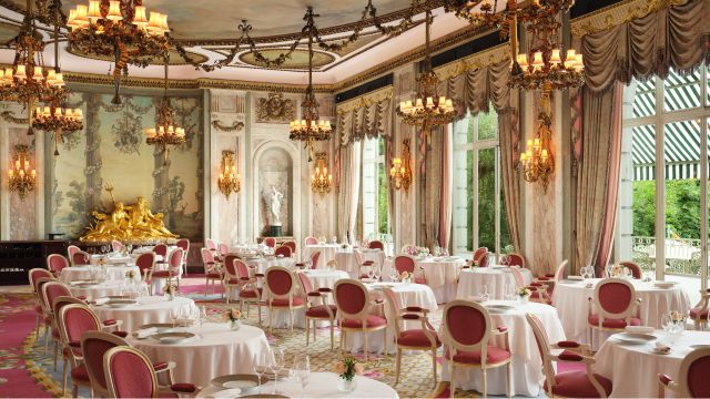 A cream chandelier lit dining room with white tables and red chairs at the ritz restaurant in london.