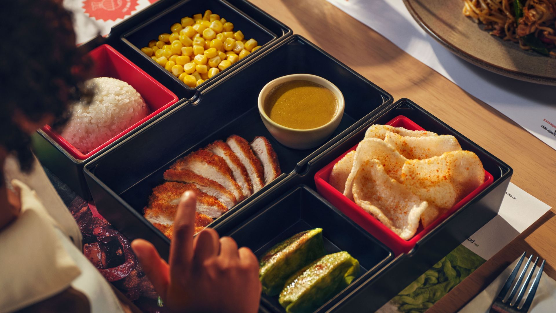 A children’s bento box meal with rice, sweetcorn, crispy chicken, dipping sauce and snacks arranged in separate compartments on a tray.