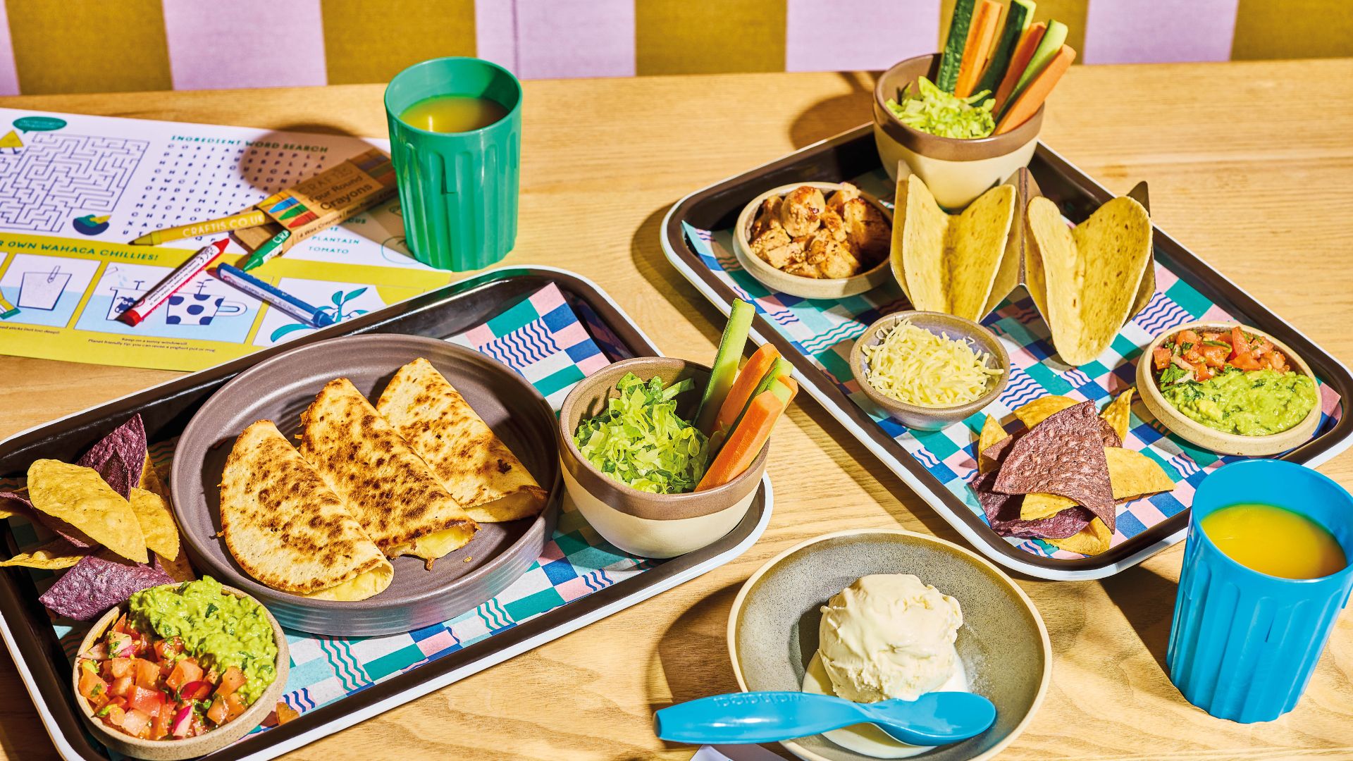 A children’s Mexican meal including quesadillas, tacos, guacamole, chips and vegetables on trays with crayons and activity sheets.