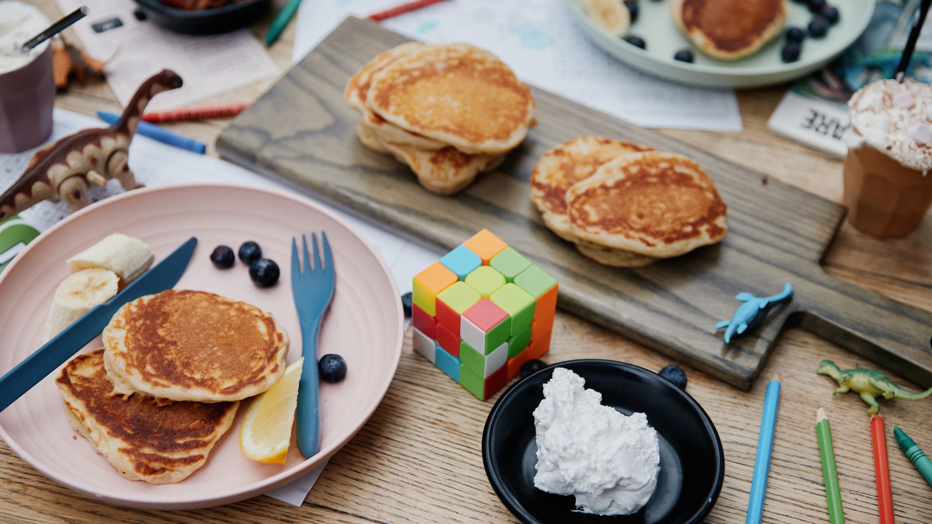A cosy breakfast scene with stacked pancakes on plates, blueberries and banana slices, a lemon wedge, and a dollop of cream, surrounded by children’s toys, crayons, and a Rubik’s cube on a wooden table.
