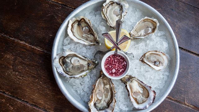 A metal tray with opened half-shell oysters with a pot of shallot vinegar and lemon, surrounded by ice.