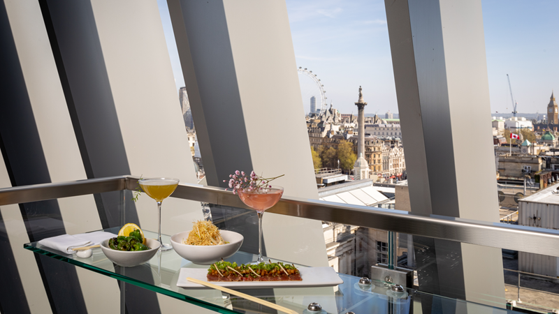 Savour a variety of creative drinks and tasty snacks with a backdrop of the city skyline. Credit: The Londoner. Image courtesy of The Londoner. Two cocktails in high glasses and three plates of Asian dishes at a glass table with a view of the London Eye and city skyline in the background from the window