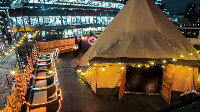 Hochwinkelaufnahme einer Rooftop-Bar mit einem mit Lichterketten geschmückten Wigwam, Feuerstellen im Freien und Sitzgelegenheiten im Freien, die an Gondeln erinnern