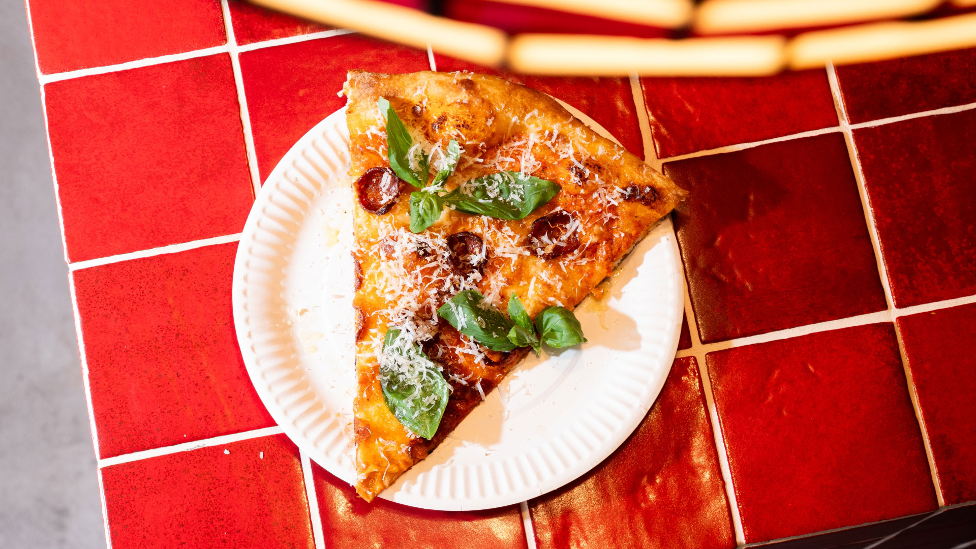 A bird's eye photo of a slice of pizza on a white paper plate on red tiles, covered in vodka sauce, pepperoni and basil leaves with a sprinkle of fresh parmesan