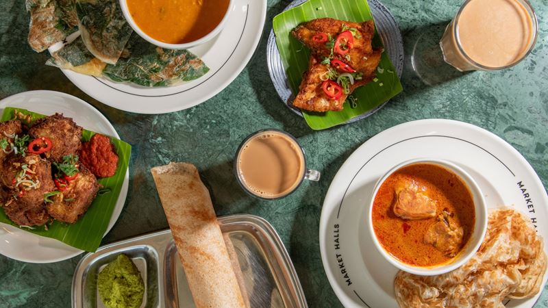 Order mouthwatering Malaysian tamil dishes from Gopal's Corner stalls across London. Credit: Pattie Tobin. Image courtesy of Gopal's Corner. A bird's-eye photo of dishes from Gopal's Corner on a table, including curries with bread, meat and vegetable sides and two drinks with Market Halls written on the plates