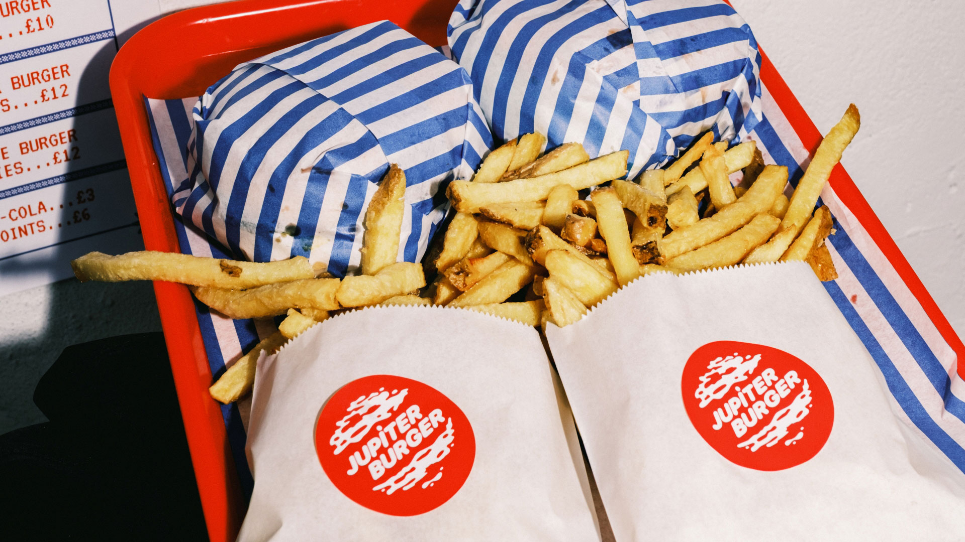 A photo of two burgers wrapped in striped paper and hand cut fries in white bags with the Jupiter Burger logo on a red tray