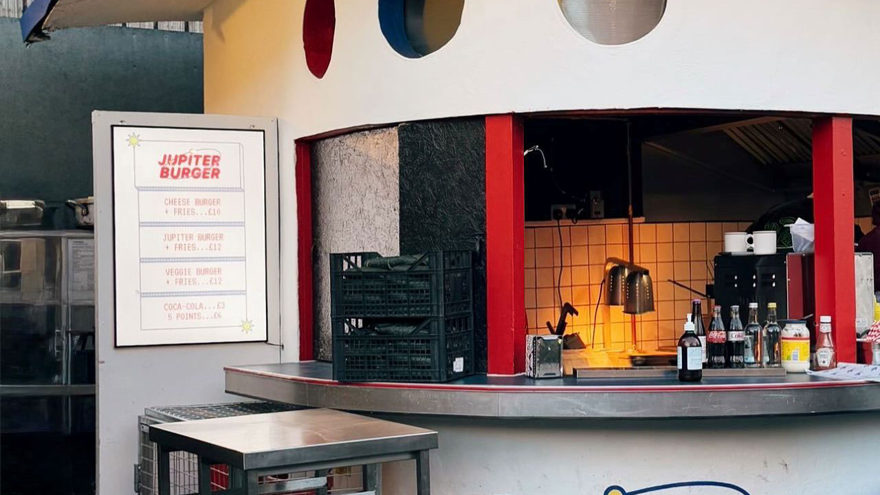 A photo of the spaceship-inspired Jupiter Burger stand at Netil Market with the menu on the wall and silver bar stools at the counter