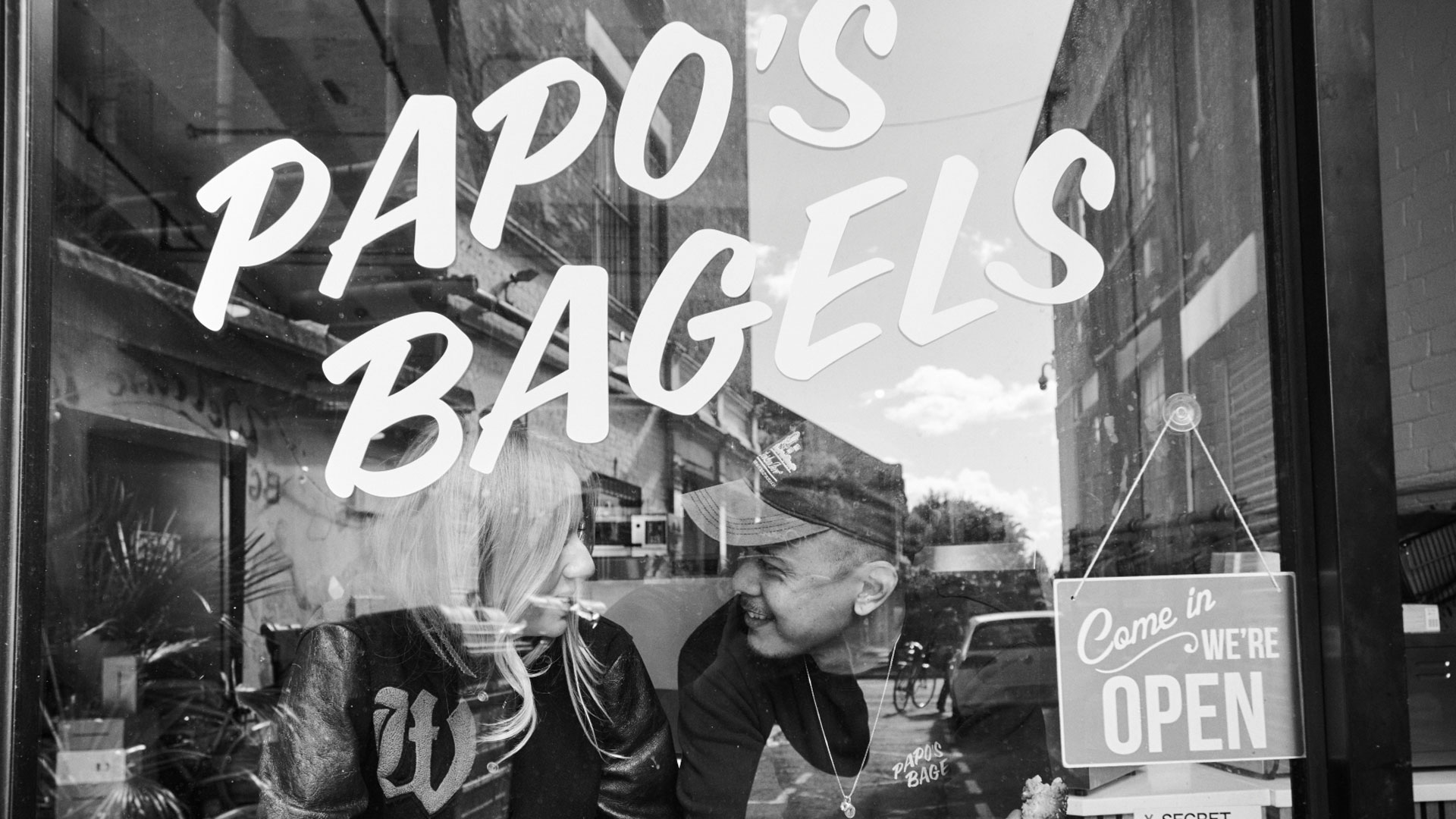 A photo of the exterior of the Papo's Bagels store with the owners in the window and a come in we're open sign
