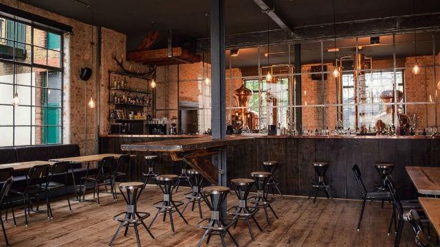 A rustic bar with brown tables and black bar stools with a low lit bar and shelves with liquor.
