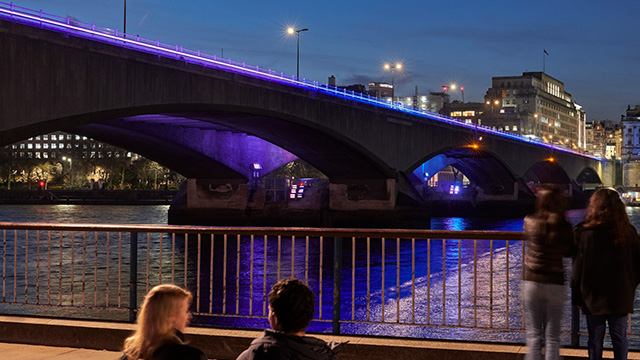 Des passants s'arrêtent le long de la Tamise pour admirer un pont illuminé de couleuir violette de nuit.