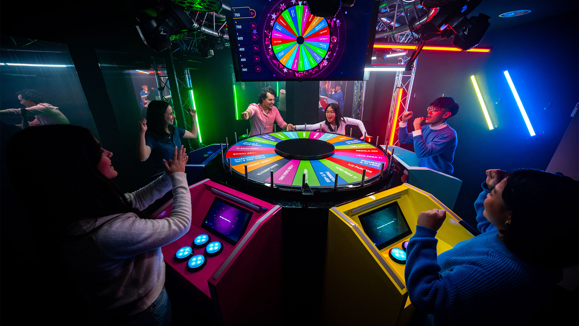 Step into the world of TV game shows with Buzz Attack, a new immersive experience complete with buzzers and games inspired by shows like Who Wants to Be a Millionaire? and Wheel of Fortune. Image courtesy of Fever. A group of people is in what looks like a game show studio, complete with a spinning wheel and individual buzzers, at the immersive London attraction, Buzz Attack!
