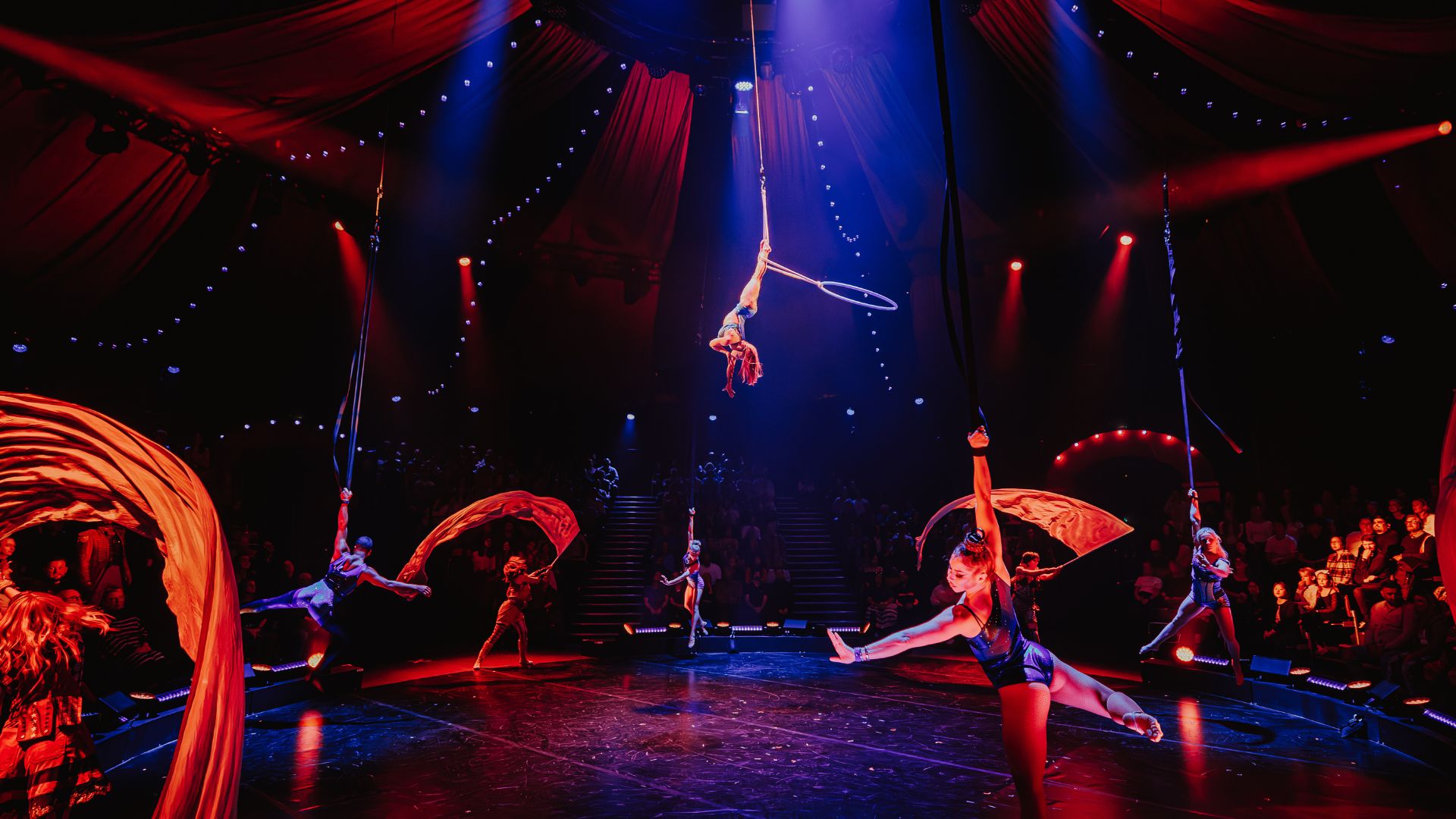 Experience the thrill of a lifetime under the big top at COME ALIVE! The Greatest Showman Circus London Spectacular. © London Theatre Direct/Luke Dyson. dark circus-themed stage with dramatic mood lighting and acrobats performing stunts