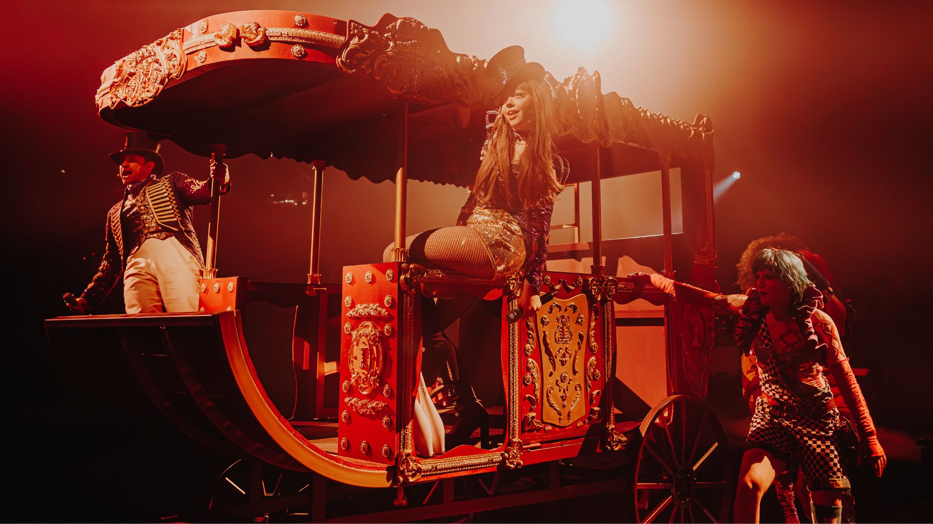 Join the greatest celebration at COME ALIVE! London: The Greatest Showman Circus London Spectacular. © London Theatre Direct/Luke Dyson three circus acts standing on a carnival cart with mood warm lighting in the background