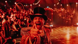 a man dressed as a magician wearing a top hat, smiling and pointing at the camera, with an audience at in the background