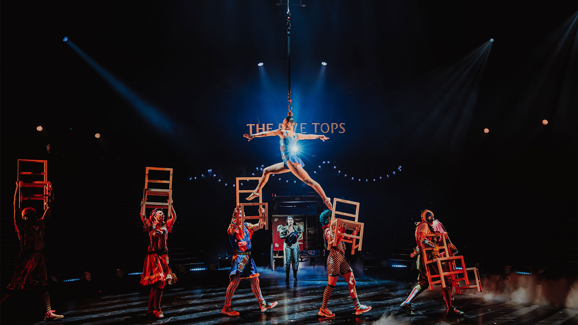 A woman wearing an electric blue bodysuit is suspended in the air by a rope tied to her hair and walking on top of elevated chairs.