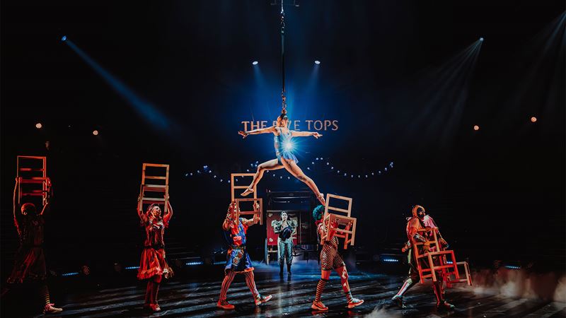 A woman wearing an electric blue bodysuit is suspended in the air by a rope tied to her hair and walking on top of elevated chairs.