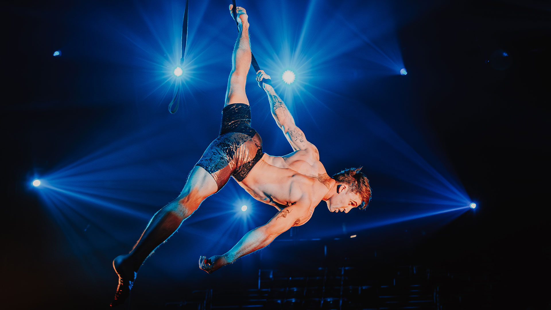 Immerse yourself in the world of circus performances and high-flying acrobatics at Come Alive! The Greatest Showman. Photo credit: Luke Dyson. An acrobat is suspended in the air by a rope held in one hand and twisted around his ankle, with blue lights in the background.
