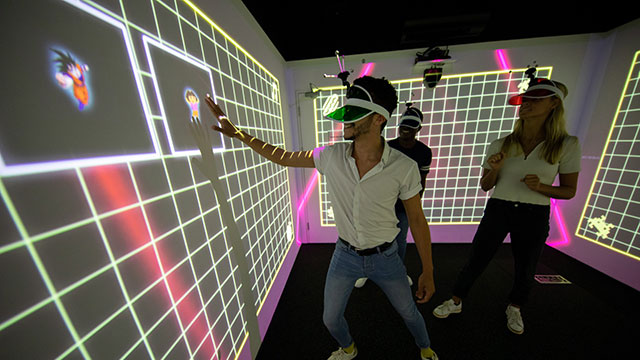 Treffen Sie sich mit Freunden bei Electric Gamebox für ein immersives Spielerlebnis © Idea Farm. Bild mit Genehmigung von Idea Farm. Eine Männer- und Frauengruppe in einer mehrfarbigen LightBox, die ein interaktives immersives Spiel spielen.