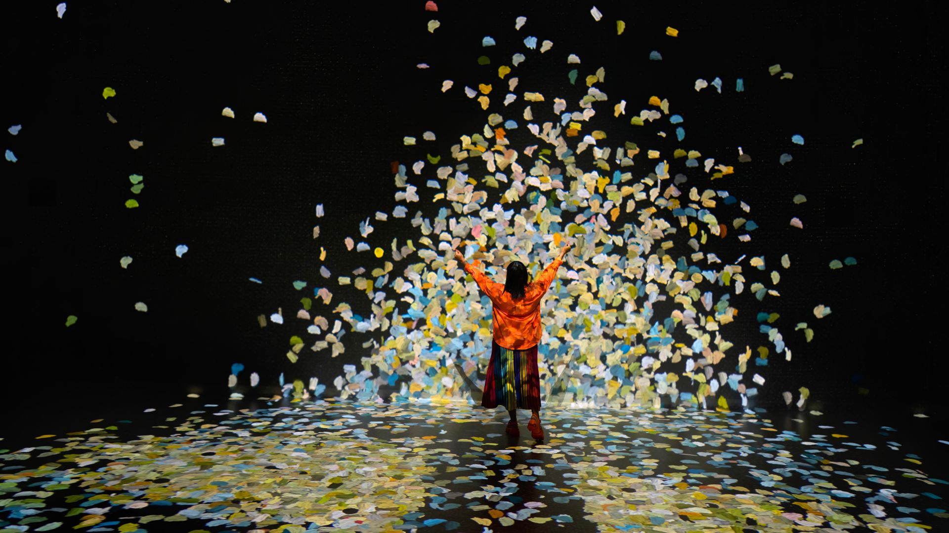 Interact with famous artworks at the epic immersive experience, FRAMELESS. Image courtesy of FRAMELESS. A woman in a red jumper and black skirt interacts with petals on a big screen at immersive art space, frameless.
