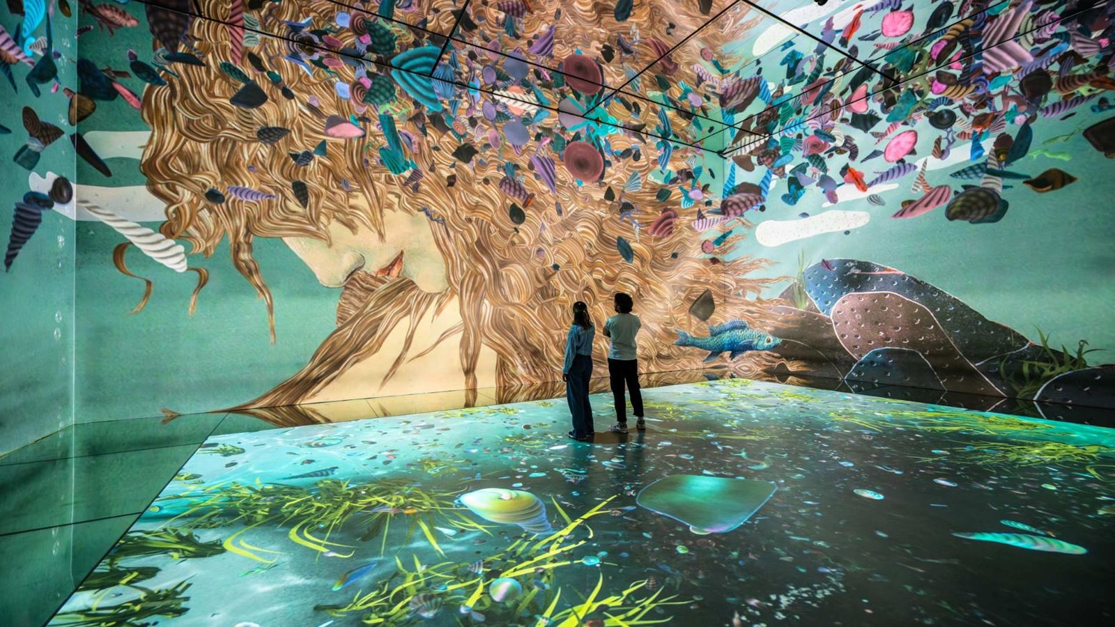 Two visitors admire a floor-to-ceiling immersive projection inspired by Botticelli's The Birth of Venus, with flowing hair, seashells and marine life surrounding the gallery.
