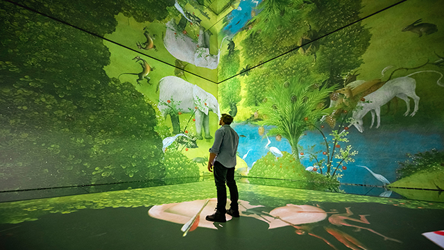The Garden of Earthly Delights in Frameless, einer immersiven Ausstellung in London. Bild mit Genehmigung von Frameless/Jordan Curtis Hughes. Ein Mann steht in einer der Galerien von Frameless und betrachtet das auf zwei Wände projizierte Gemälde „Der Garten der Lüste".