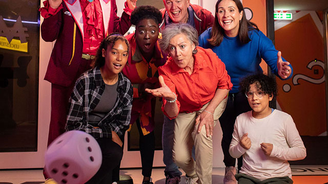 Erleben Sie in diesem Sommer spannende Spiele und epische Herausforderungen bei Monopoly Lifesized © Helen May Banks. Bild mit Genehmigung von Borkowski Arts & Ents. Eine Frau wirft einen großen Würfel vor einer Gruppe von Kindern und Erwachsenen.