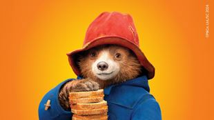 Paddington bear in a blue coat and red hat holding a stack of marmalade sandwhiches.