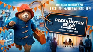 Paddington bear in a blue coat and red hat, running holding a brown suitcase with bunting behind him