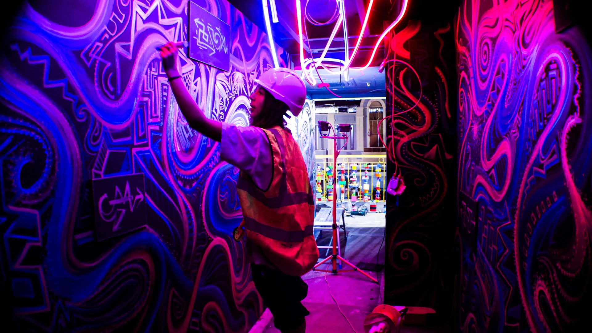 A person wearing a hard hat and high-visibility vest explores a vividly painted neon corridor inside Wake The Tiger's immersive experience, surrounded by glowing artwork and colourful lighting.