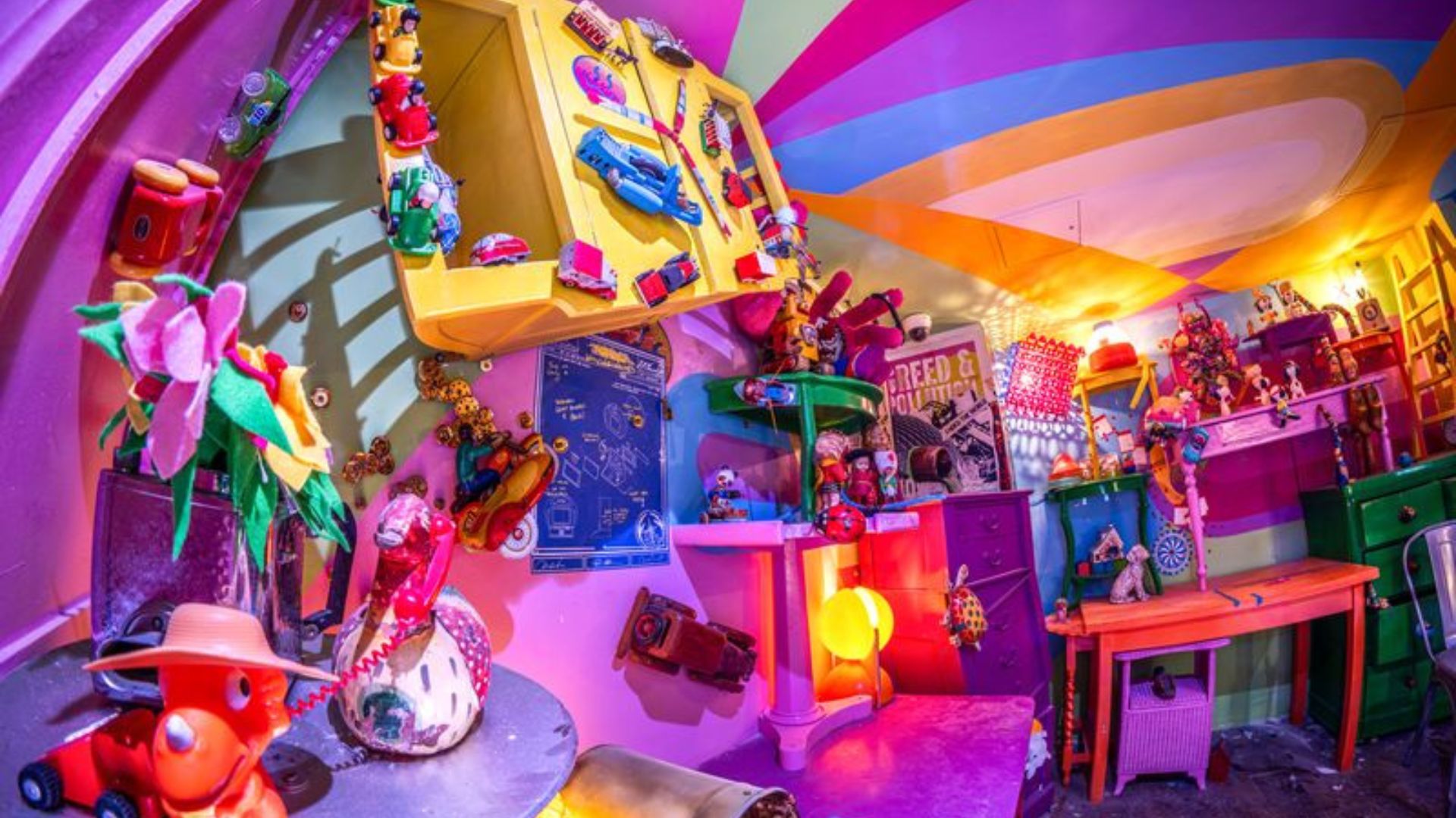 From rainbow ceilings to toy-filled walls, every corner of Absurd City is packed with unexpected details. Keep your eyes peeled – you never know what you might find around the next corner. Image courtesy of We Are Plaster. Photo credit: Andre Pattenden A brightly coloured room inside Wake The Tiger's immersive experience, filled with toys, artwork, colourful furniture and quirky objects beneath a rainbow-striped ceiling.