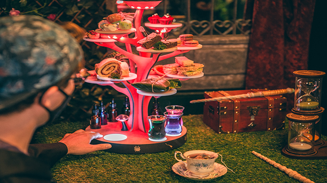 Wizard Afternoon Tea beim The Wands & Wizard Exploratorium. Bild mit Genehmigung von The Cauldron Ein Mann berührt den Nachmittagstee-Stand in Form eines rosa beleuchteten Baumes, auf dem eine Vielzahl von Kuchen und eine Auswahl von Molekulargetränken stehen.