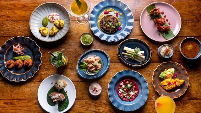 A collection of colourful small plates on a wooden table and viewed from above, featuring Indian food, from starters to desserts and two cocktails.