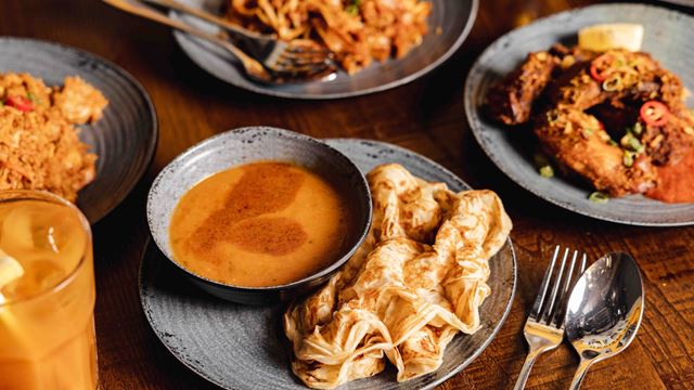Three Malaysian dishes laid out on a wooden table, including a flaky roti with sauce.