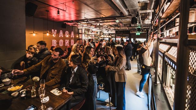 Crowds of people chatting and enjoying drinks inside Vagabond wine bar with Vagabond letters spelt out in pink neon lights on the wall.