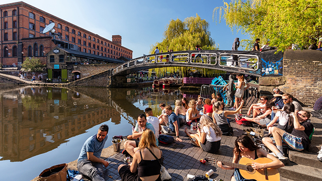 Camden Lock © visitlondon.com / Jon Reid. Image courtesy of visitlondon.com / Jon Reid. People sit beside Camden Lock on a sunny day, some chatting, some drinking and one playing a guitar - with a bridge and the canal in the background.