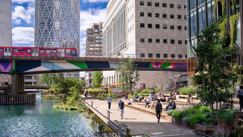 Eden Park in Canary Wharf with DLR train on a bridge and people walking by the river below.