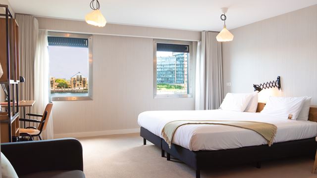Double hotel room with bed in white sheets, white walls, carpeted floors and windows with views of Royal Victoria Dock.