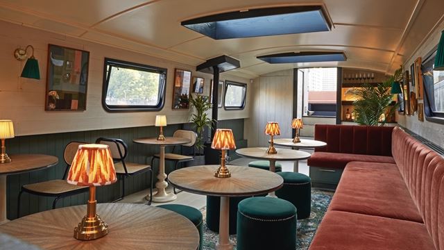 Inside The Boathouse cocktail bar, featuring retro lamps, pink and green velvet seats and banquette and pictures hung on the boat's walls.