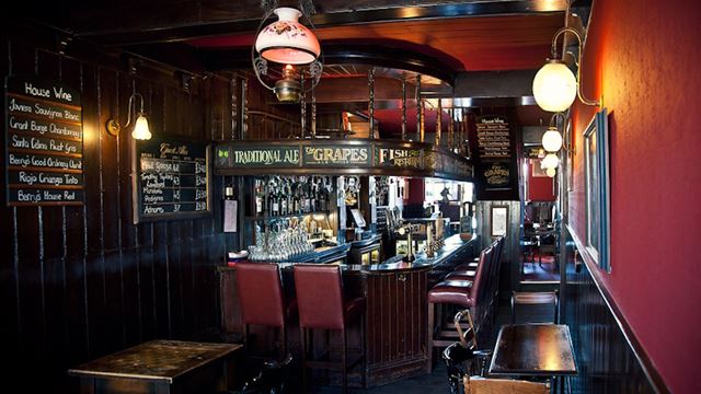 Darkly lit interior of The Grapes pub with red and dark brown walls, dark brown bar counter and vintage lamps.