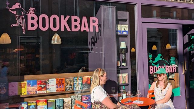 Two people sit down to enjoy a glass of wine on a table outside BookBar, with the shop window in the background and books on display.