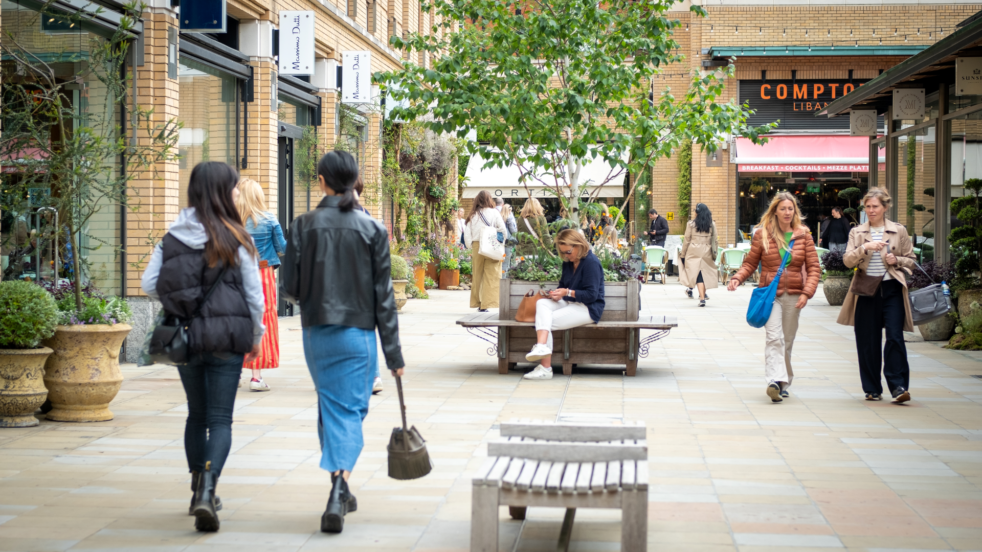 Two women walk away from the camera at Duke of York Square with people in the distance, benches, trees and shops and restaurants in the background