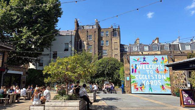 A view of Eccleston Yards' courtyard, featuring a colourful mural saying "Welcome to Eccleston Yards" and benches with people sitting on them.