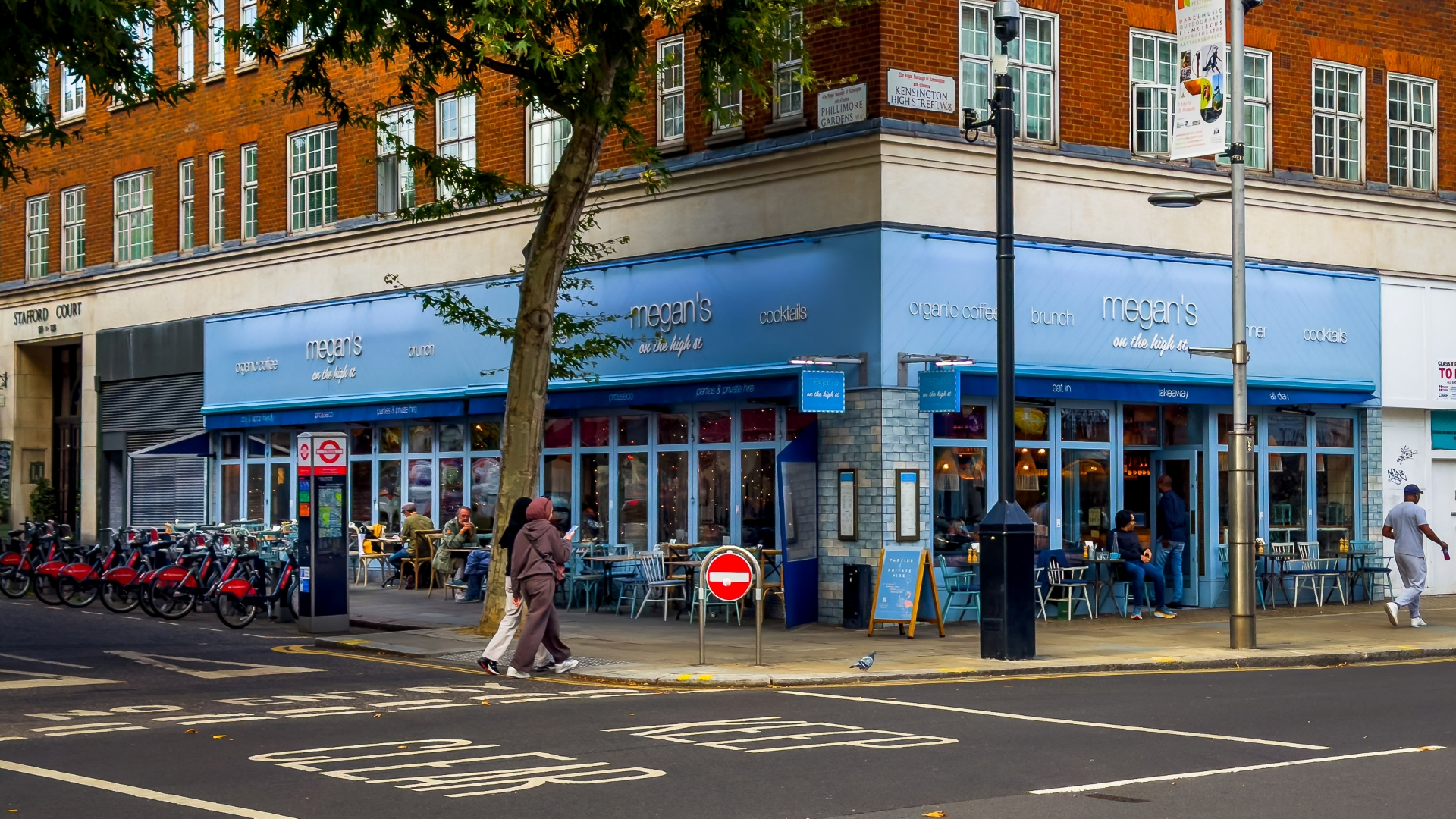 A photo of Megan's Cafe in Chelsea from outside, a light blue building on a corner with people walking outside and a Santander docking station