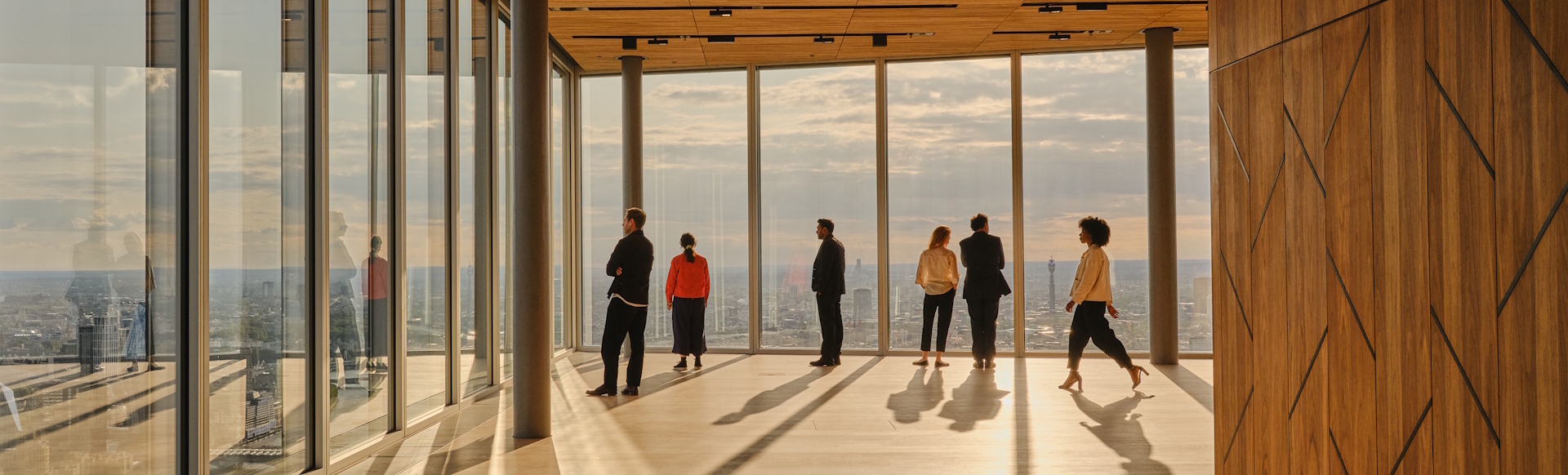 A group of people look at the view of London from a viewing platform on the 50th floor of a building with glass floor-to-ceiling windows, as the sun sets.