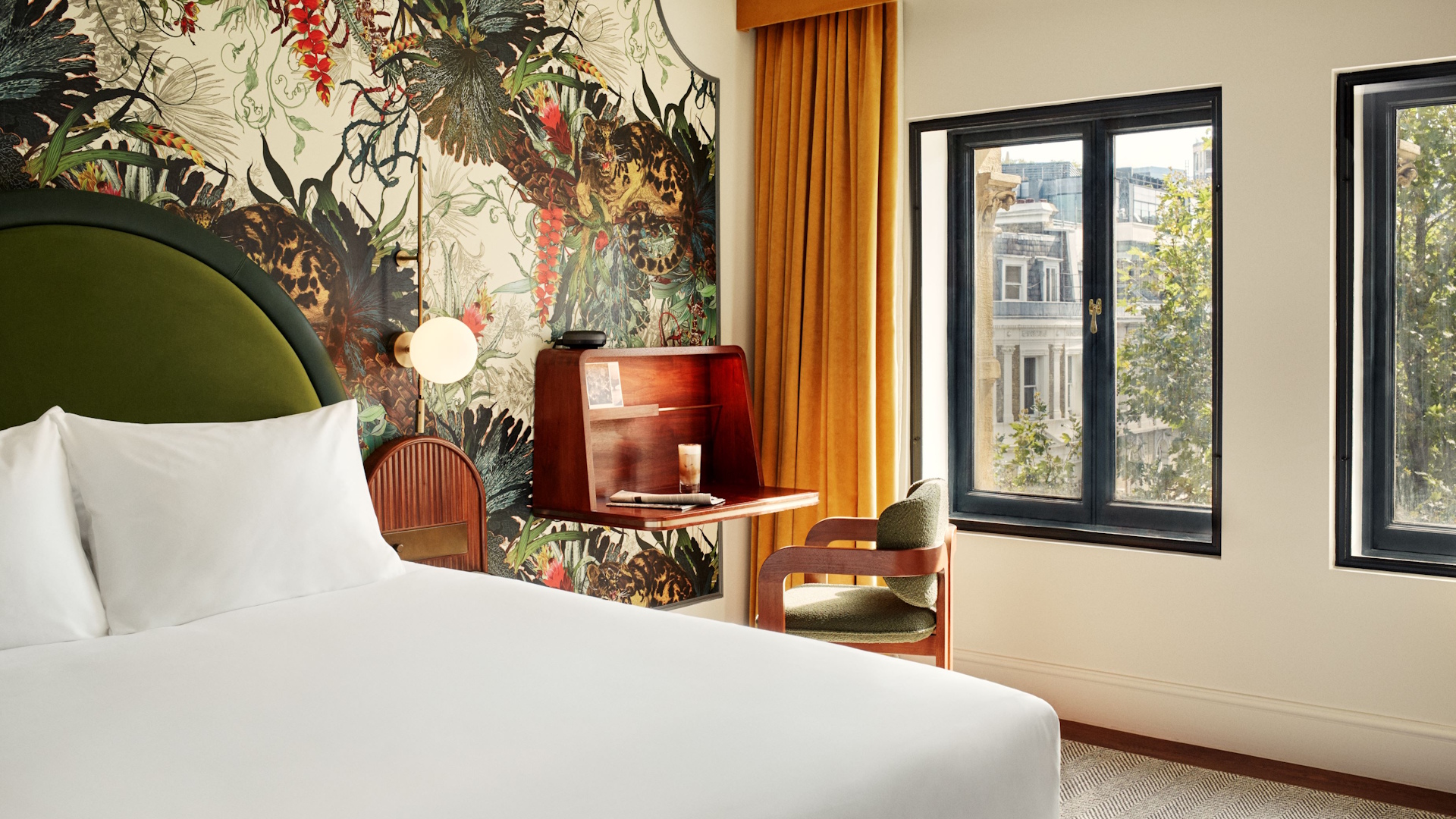 Hotel bedroom with double bed with white sheets, green and red wallpaper, orange curtains and wooden furniture.
